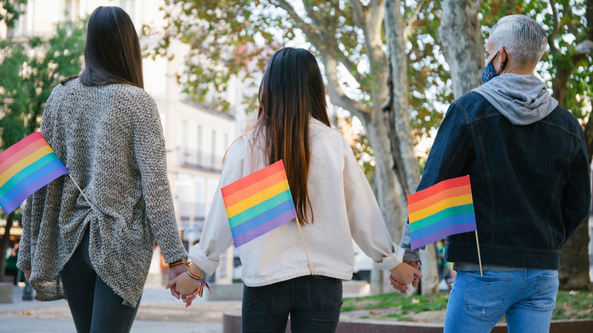 Drie mensen hand in hand op straat, op de rug gezien met LGBTIQ+-vlaggetje in hun achterzak.