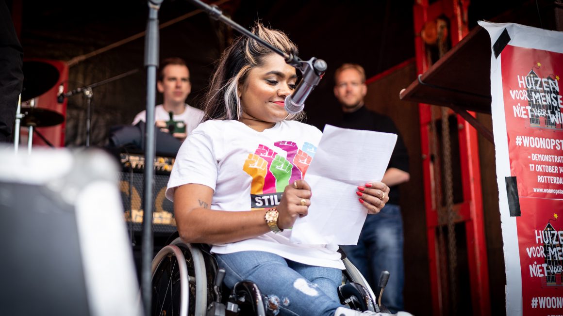 Een foto van Jeanette Chedda, enigszins van onderen genomen terwijl zij in haar rolstoel voor een microfoon zit. Ze draagt blauwe jeans en een wit T-shirt met kleurrijke opgestoken vuisten. Ze leest voor van een A4-papier.