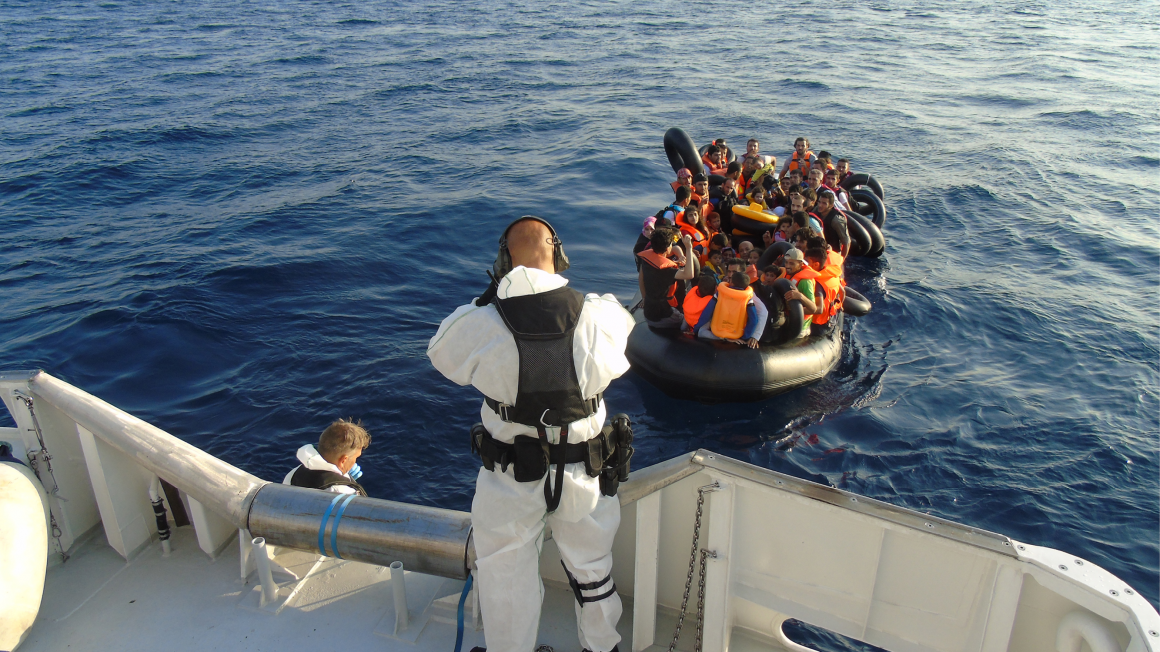 Twee personen in witte pakken staan op een hoge witte boot. Ze staan met hun rug naar de camera en ze kijken naar een klein zwart rubberbootje vol met mensen. Deze mensen hebben allemaal reddingsvesten aan en zwembanden vast.